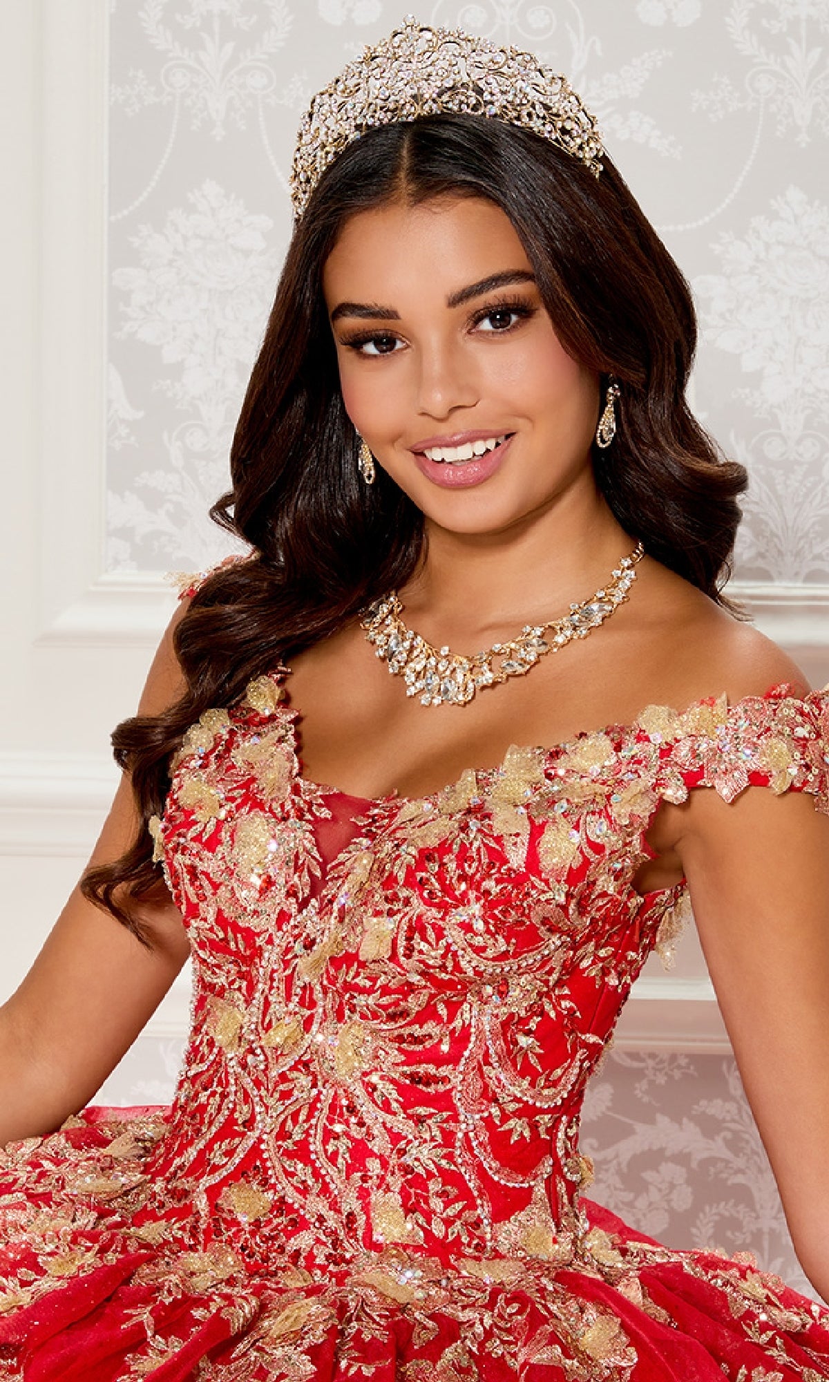 A young woman poses in front of a patterned white wall, wearing the Quinceanera Dress PR30119 By Princesa—an elegant red and gold embroidered off-the-shoulder gown with a jeweled tiara and necklace.